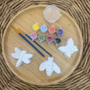 Peut inclure: Un kit de loisirs créatifs comprenant trois figurines en plâtre blanc en forme de libellule, d'abeille et de papillon. L'ensemble inclut deux pinceaux à manche bleu, une bande de huit petits pots de peinture et un récipient en plastique vide.