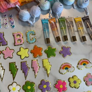 May include: A collection of colorful accessories and beauty products. Includes plush headbands, lip gloss tubes, decorative patches in various shapes like stars, flowers, hearts, lightning bolts, and rainbows. The patches have gold trim.