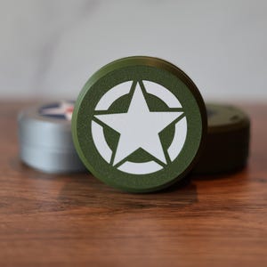 May include: A green, round container with a white star emblem inside a white circle. The container is on a wooden surface, with two other containers slightly out of focus in the background.
