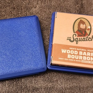 May include: A blue soap bar container with a brown soap bar inside. The soap bar is labeled "Dr. Squatch Wood Barrel Bourbon Men's Natural Soap Jimmy Red Corn & Sand Medium Grit 5 oz Bar".