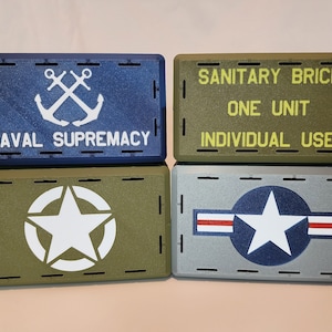 May include: Four small, rectangular boxes stacked in two rows. The top row has a blue box with a white anchor and the text "NAVAL SUPREMACY" and a green box with the text "SANITARY BRICK ONE UNIT INDIVIDUAL USE" in yellow. The bottom row has a green box with a white star in a circle and a gray box with a white star and red stripes.