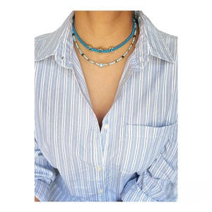 May include: A layered necklace with three strands of turquoise blue beads and gold accents. The necklace is worn by a person with a striped shirt.