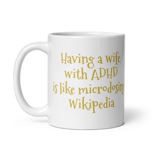 May include: White ceramic coffee mug with a gold inscription that reads "Having a wife with ADHD is like microdosing Wikipedia".