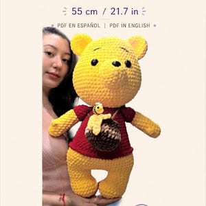 May include: A crochet pattern for a Winnie the Pooh plush toy, measuring 21.7 inches. The yellow bear wears a red shirt and carries a brown honey pot with a small bee. The document includes text for beginner to intermediate skill levels.