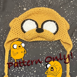 May include: A crocheted yellow hat with a dog face, white eyes, and a black nose. The hat has long, braided tails. The text "Pattern Only!" is written in red on a gray background with white dots.