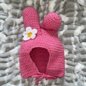 以下が含まれることがあります： ピンクの毛糸で編まれたウサギの帽子。側面に白い花が飾られています。帽子にはフードとリボンが付いています。