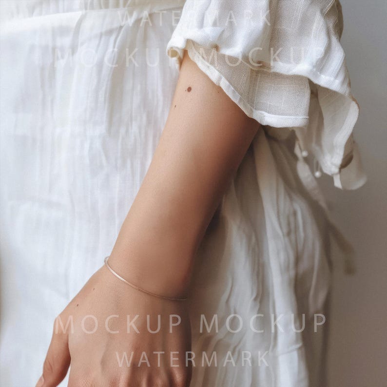 K&ouml;nnte beinhalten: Ein zartes, d&uuml;nnes Silberarmband schm&uuml;ckt das Handgelenk einer Person. Das Armband hat ein schlichtes, elegantes Design. Die Person tr&auml;gt ein wei&szlig;es Kleid mit R&uuml;schen&auml;rmeln. Das Bild ist eine Nahaufnahme.