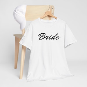 Puede incluir: Una camiseta blanca con la palabra "Bride" en escritura negra. La camisa está colgada en una percha de madera. Una gorra de béisbol blanca descansa sobre el respaldo de una silla de madera.
