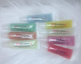 May include: An assortment of lip gloss tubes in various colors, including blue, green, yellow, peach, pink, and red. Each tube has a clear applicator and is labeled with "DM Creations". The background is a soft, white, fluffy texture.
