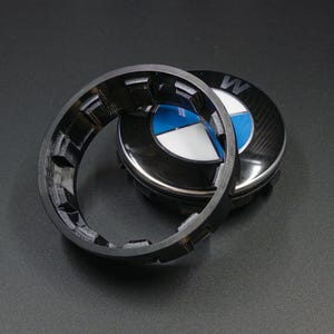 Könnte beinhalten: Eine schwarze Radnabenabdeckung mit dem BMW-Logo in Blau, Weiß und Schwarz. Ein schwarzer, strukturierter Ring befindet sich vor der Kappe. Der Hintergrund ist eine dunkelgraue Oberfläche.