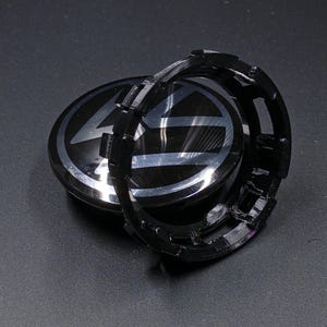May include: Black car wheel center cap with a silver emblem. The cap has a glossy finish and a black plastic retaining ring. The emblem features a stylized design on a black background.
