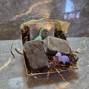 May include: A gift box containing three dark-colored soap bars and two toy dinosaurs. The soap bars are rectangular and wrapped in clear plastic. The dinosaurs are green and purple. The box is filled with brown paper shreds.