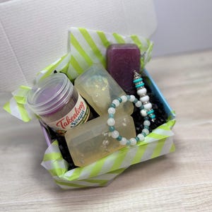 May include: A gift box filled with a variety of self-care items. The box contains a jar of Takealong Soaps, two bars of soap, a purple bar of soap, and a bracelet with turquoise and white beads. The box is lined with green and white striped tissue paper.