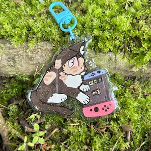 Peut inclure: Un porte-clés transparent avec un chat de dessin animé tenant une Nintendo Switch rose. Le chat a du pelage brun, blanc et orange et est entouré de paillettes.