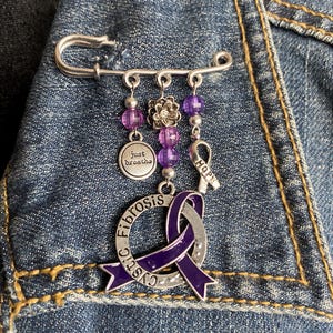 May include: A silver safety pin brooch with dangling charms. The charms include a purple ribbon with the words "cystic fibrosis", a small silver disc with "just breathe", a flower, purple beads, and a ribbon with the word "hope". The brooch is pinned to a denim jacket.