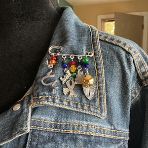 May include: A silver safety pin adorned with charms and colorful beads. Charms include a snake, ankh, building, and a gold-colored feather. The pin is attached to a denim jacket.