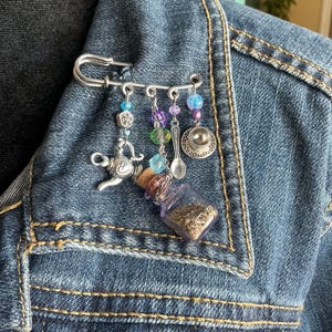 May include: A decorative safety pin brooch with dangling charms. The charms include a miniature teapot, a small bottle with a cork, a spoon, a teacup, and colorful beads. The brooch is attached to a denim jacket.