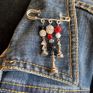 May include: A silver safety pin brooch with dangling chess-themed charms. The charms include a silver knight and two chess pieces, along with decorative beads in red, white, black, and clear crystal.
