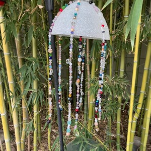 May include: A decorative outdoor solar-powered light fixture with a silver-colored, half-dome shade. Colorful beaded strands hang from the shade. The light is suspended in front of a bamboo background, with a large green glass sphere at the bottom.