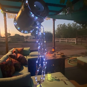 May include: A decorative outdoor water feature resembling a metal pitcher pouring a stream of lights. The pitcher is suspended from a wooden structure, with the lights cascading down. The scene is set outdoors with seating and a sunset in the background.