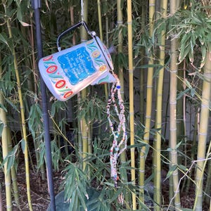May include: A decorative metal watering can with a black handle and spout, painted with a floral design in red, white, and blue. The can appears to be pouring out a stream of colorful beads. The text on the can reads "Raffletarch är den bästa av alla jordiska drycker."