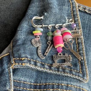 May include: A silver-tone sewing-themed brooch pinned to a denim jacket. The brooch features charms of a tape measure, clothespin, scissors, and a Singer sewing machine. Colorful beads and pink thread spools add detail.