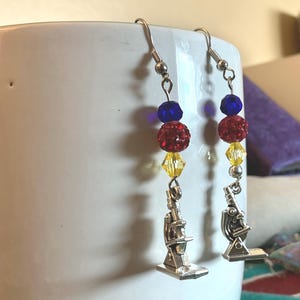 May include: A pair of dangle earrings with a science theme. Each earring features a silver-colored microscope charm, red and blue beads, and yellow crystal beads. The earrings hang from silver-colored fish hook ear wires.