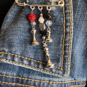 May include: A silver safety pin with three dangling charms. The charms include a red beaded sphere, a white gem, and a black beaded charm. Each charm has a silver chess piece at the bottom. The pin is attached to a blue denim jacket.
