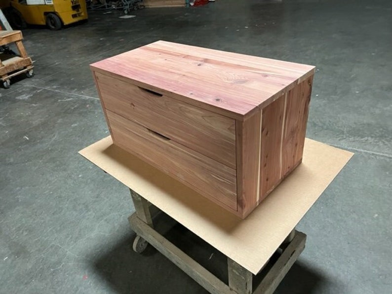May include: A two-drawer wooden chest with a natural wood finish. The chest has two drawers with simple metal handles. The chest is sitting on a light brown surface.