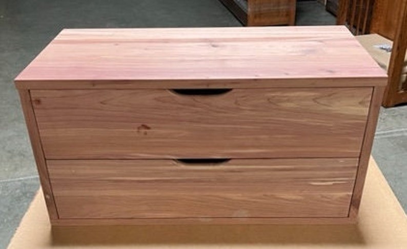 May include: A two-drawer wooden chest with a natural wood finish. The chest has simple, recessed handles.