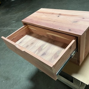 May include: A wooden drawer unit with a single drawer that is open. The drawer is made of light brown wood and has a smooth finish. The drawer unit is sitting on a brown surface.