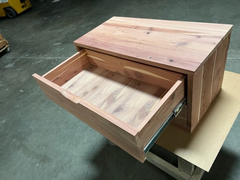 May include: A wooden drawer unit with a single drawer that is open. The drawer is made of light brown wood and has a smooth finish. The drawer unit is sitting on a brown surface.