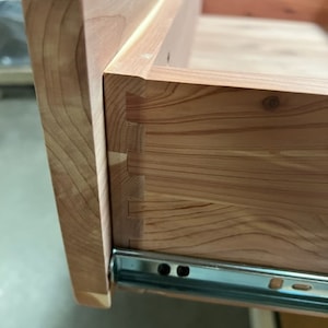 May include: Close-up of a wooden drawer with a dovetail joint and a metal drawer slide. The drawer is made of light-colored wood with a natural grain pattern.