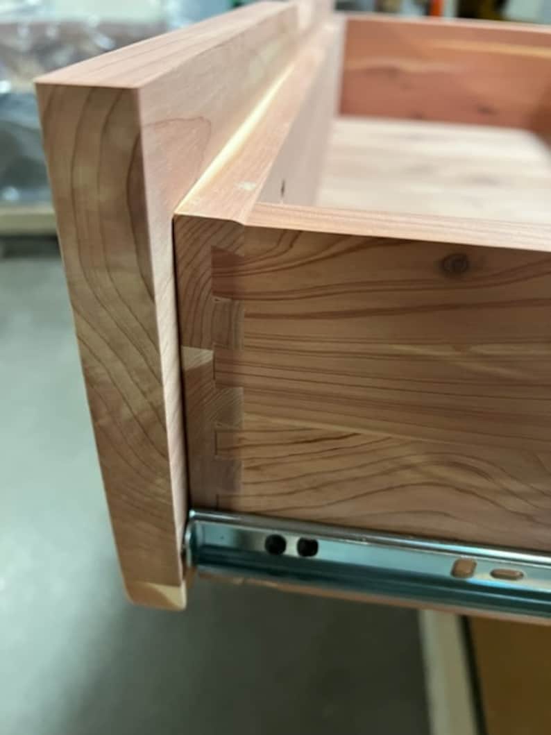 May include: Close-up of a wooden drawer with a dovetail joint and a metal drawer slide. The drawer is made of light-colored wood with a natural grain pattern.