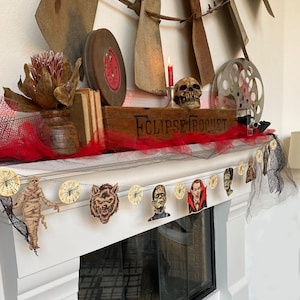 May include: A Halloween-themed garland with a black and white spiderweb design. The garland features various characters, including a werewolf, a vampire, a Frankenstein monster, and a mummy. The garland is hung on a white fireplace mantel with a red and black tulle backdrop.
