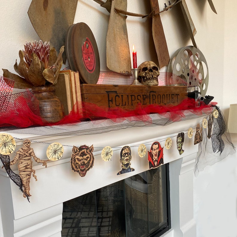 May include: A Halloween-themed garland with a black and white spiderweb design. The garland features various characters, including a werewolf, a vampire, a Frankenstein monster, and a mummy. The garland is hung on a white fireplace mantel with a red and black tulle backdrop.