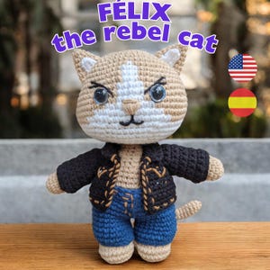 Può includere: Un gatto all'uncinetto che indossa una giacca di pelle nera e jeans blu. Il gatto ha una faccia bianca con occhi neri e un naso nero. Il gatto è in piedi su una superficie di legno. Il testo "FÉLIX the rebel cat" è scritto sopra il gatto. Le bandiere americana e spagnola sono nell'angolo in alto a destra dell'immagine.