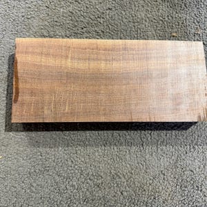 May include: A rectangular block of natural wood with a smooth, flat surface. The wood features a medium brown colour with visible horizontal grain patterns and subtle darker streaks running across the length of the piece.