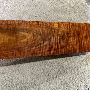 May include: A rectangular block of wood featuring a rich, warm amber and brown colour palette. The surface displays a prominent, wavy, and shimmering tiger-stripe grain pattern that runs horizontally across the length of the wooden piece.