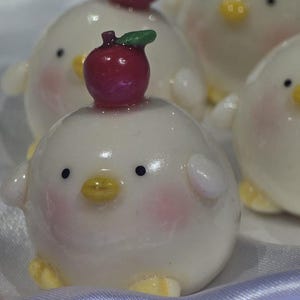 May include: A collection of white, round chick figurines with rosy cheeks, black eyes, and yellow beaks. Each chick has a red apple with a green leaf on its head. The figurines are on a white, silky surface.