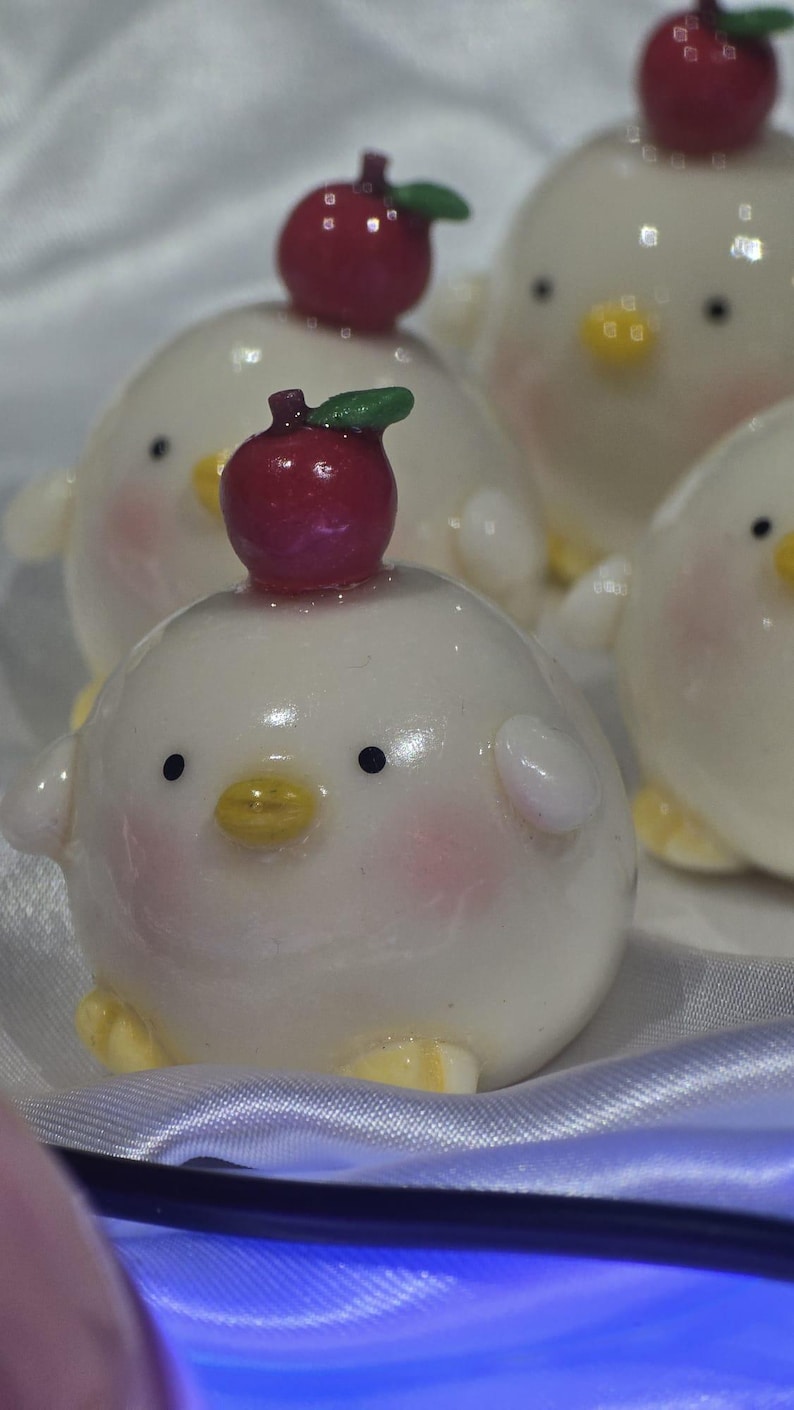 May include: A collection of white, round chick figurines with rosy cheeks, black eyes, and yellow beaks. Each chick has a red apple with a green leaf on its head. The figurines are on a white, silky surface.