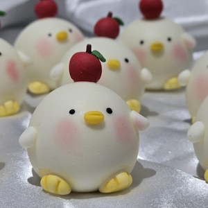 May include: A collection of adorable, round, white chick-shaped treats, each topped with a red apple. The chicks have pink cheeks, black eyes, yellow beaks, and yellow feet. The treats are arranged on a white surface.