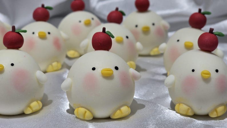 May include: A collection of adorable, round, white chick-shaped treats, each topped with a red apple. The chicks have pink cheeks, black eyes, yellow beaks, and yellow feet. The treats are arranged on a white surface.