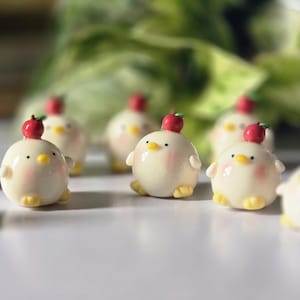 May include: A collection of small, round, cream-colored chick figurines with rosy cheeks, yellow feet, and tiny red apple hats. The figurines are arranged on a white surface, with a blurred green background.