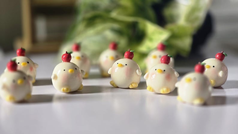 May include: A collection of small, round, cream-colored chick figurines with rosy cheeks, yellow feet, and tiny red apple hats. The figurines are arranged on a white surface, with a blurred green background.