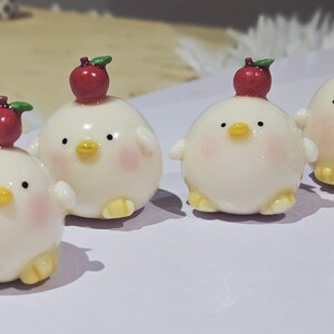 May include: Four adorable, round, white chick figurines with small wings and yellow feet. Each chick has a red apple on its head and rosy cheeks. The figurines are arranged on a light-colored surface.