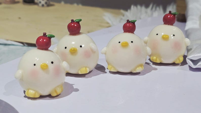 May include: Four adorable, round, white chick figurines with small wings and yellow feet. Each chick has a red apple on its head and rosy cheeks. The figurines are arranged on a light-colored surface.