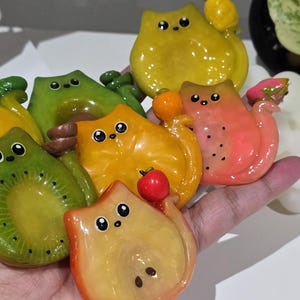 Può includere: Una collezione di figurine di gatti colorati a forma di frutta. Le figurine hanno la forma di vari frutti, tra cui kiwi, mela, mango e pesca. Ogni gatto ha una faccia carina ed è dipinto con colori vivaci. Le figurine sono tenute in una mano.