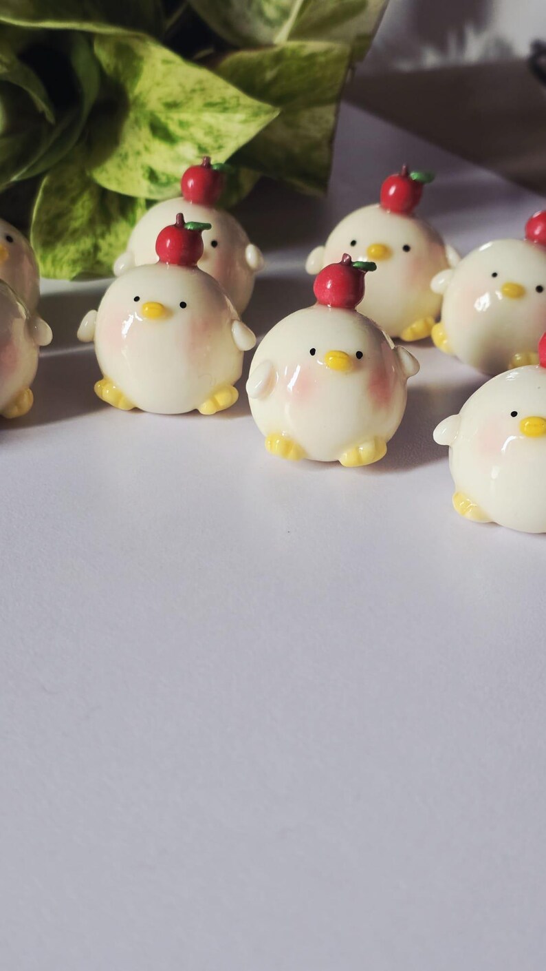 May include: A collection of small, round, white figurines resembling chicks, each with a red apple on its head. The chicks have yellow feet, small black eyes, and pink cheeks. The background is a soft white surface.