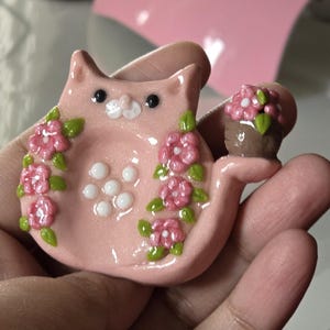 May include: A pink cat-shaped trinket dish with a small flower pot attached. The dish is decorated with pink flowers and green leaves. The cat has black eyes and white details. The trinket dish is held in a hand.